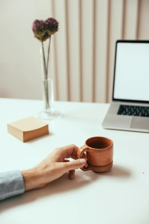 a person holding onto a small mug with a flower in a vase nearby