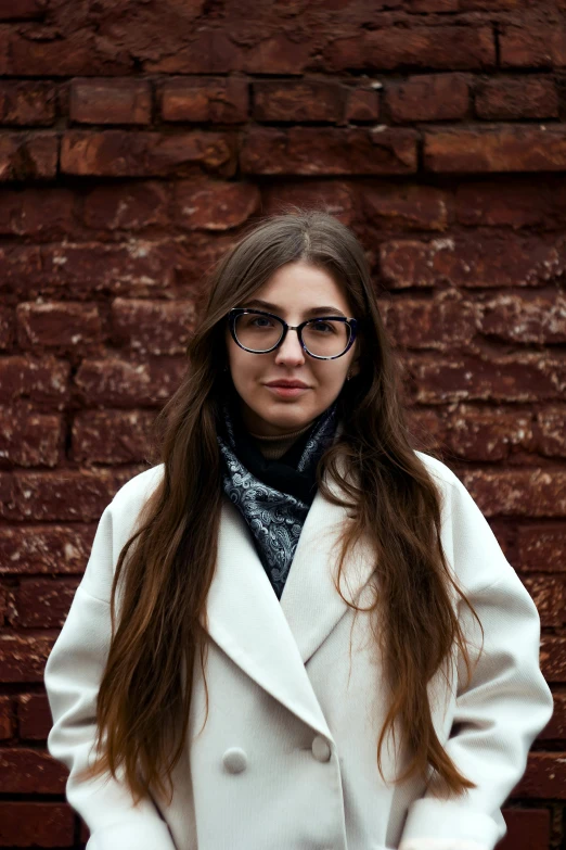 a girl with glasses and a pea coat stands against a brick wall