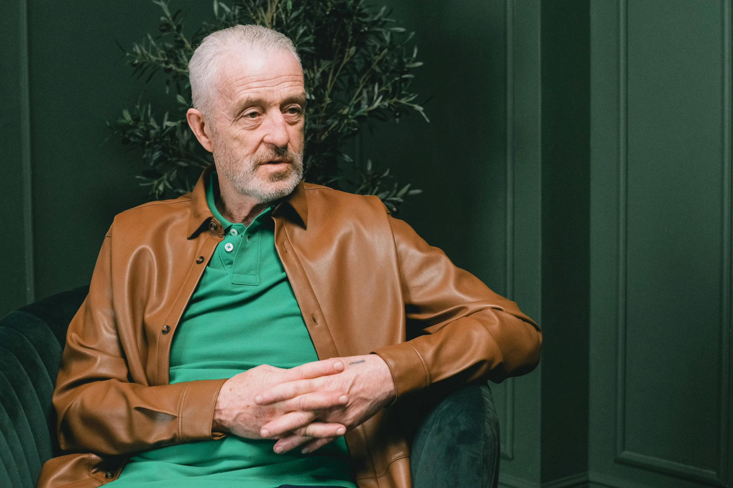 a bald man with white hair and a green shirt, sitting in a chair wearing a leather jacket