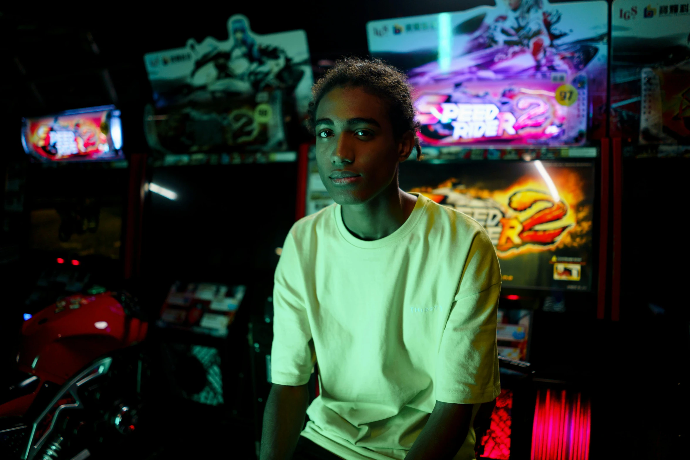 young man in white shirt looking at camera while sitting in front of neon video game machines