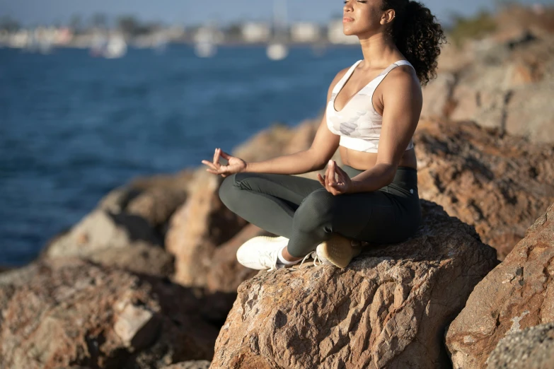 a woman in yoga gear meditates on a rock next to the water