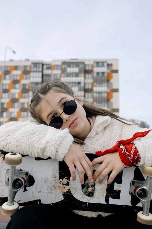 a girl with sunglasses and coat sits on top of a skateboard