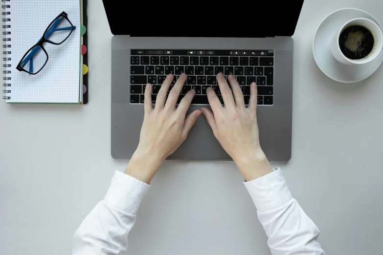 hands on the keyboard of a laptop with a pen, mug and coffee cup