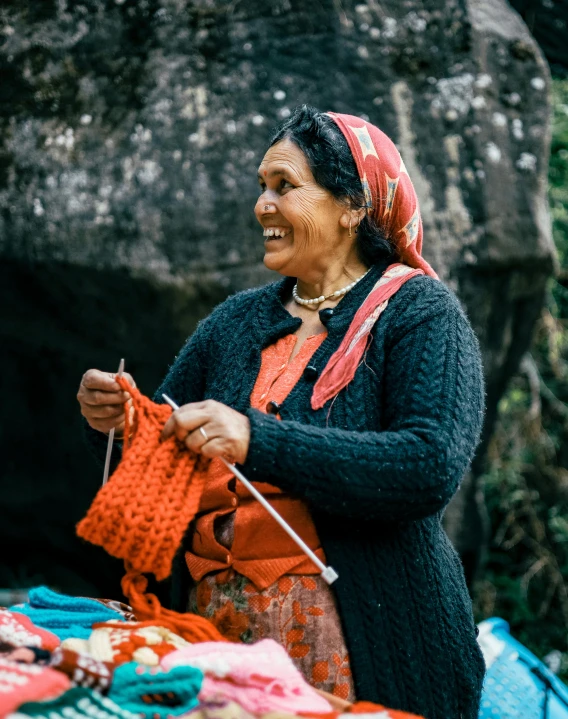 the woman smiles as she holds a red crochet hook near a pile of clothing