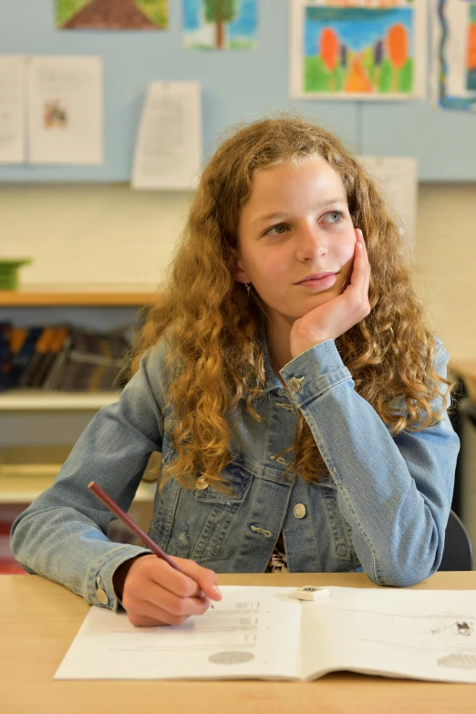 a girl with long hair in a denim jacket is sitting at a desk