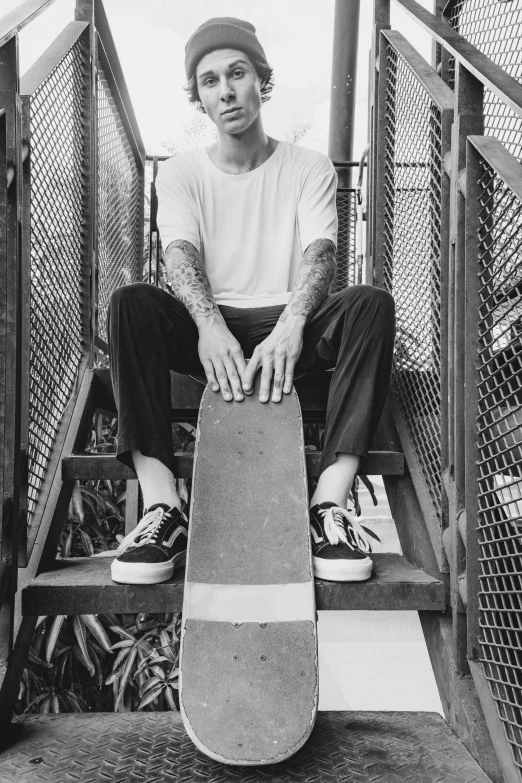 a young man with glasses, hat, and beard sits on stairs while holding a skateboard