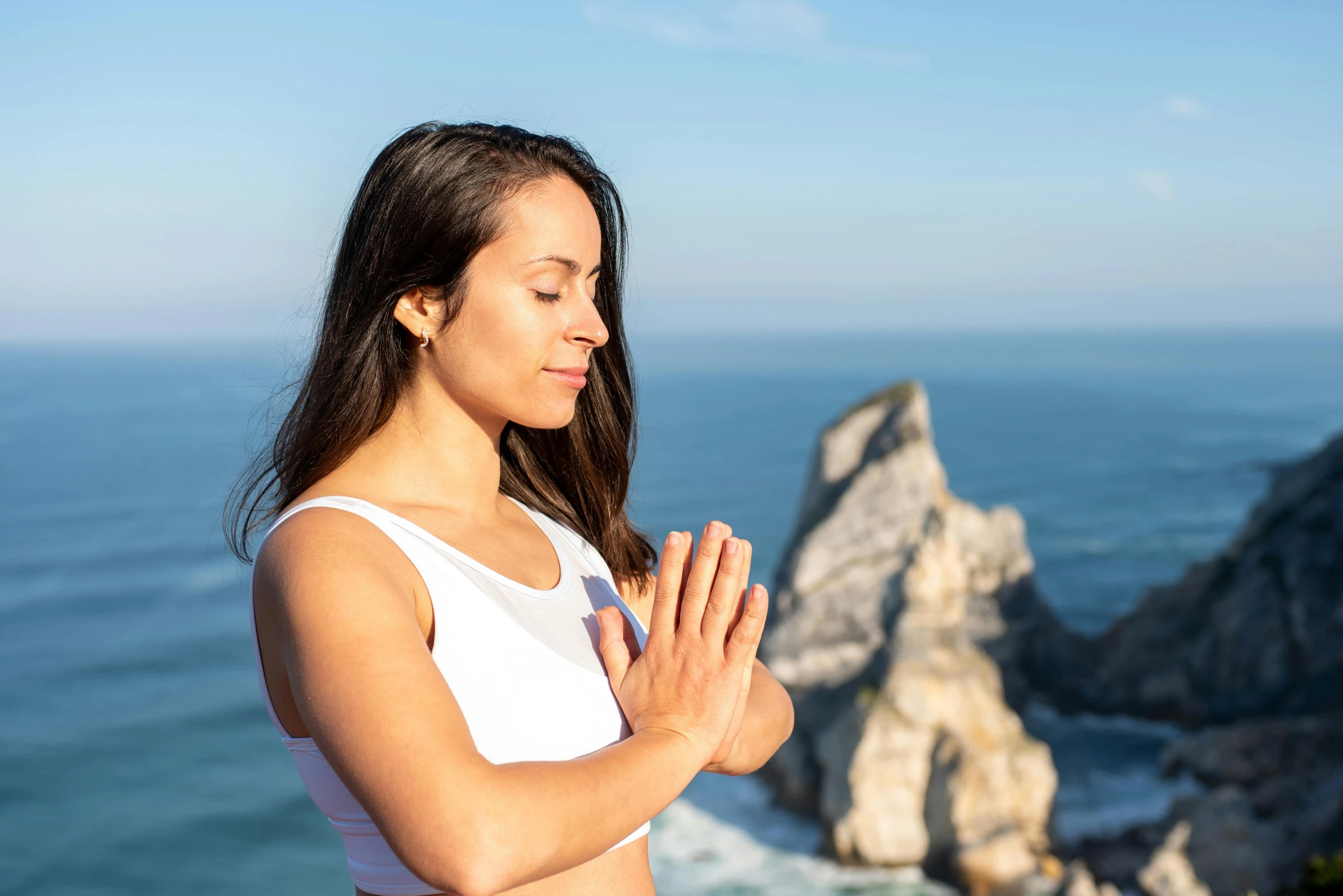 woman standing in front of the ocean in a white top and wearing her hands together