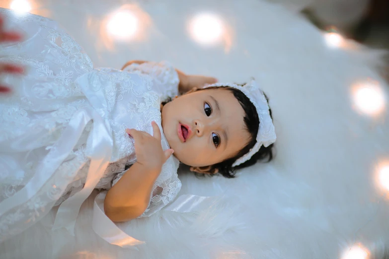 an adorable child with a white dress laying on top of a fluffy white sheet