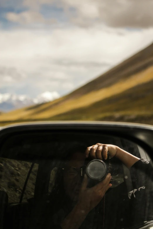 a person holding a lens in their hand is reflected in the side mirror of a vehicle