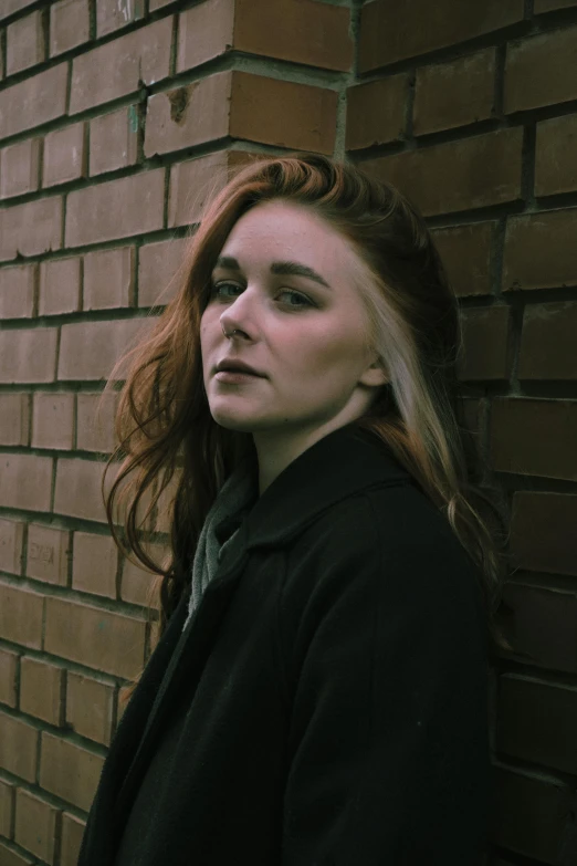 a woman with red hair standing against a brick wall