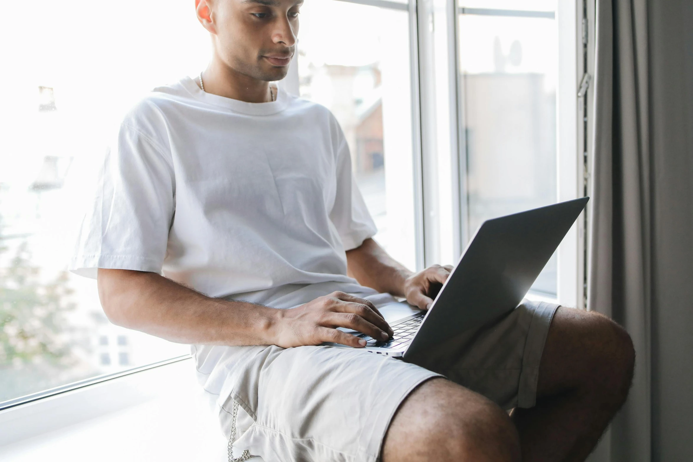 a man sitting near a window typing on his laptop