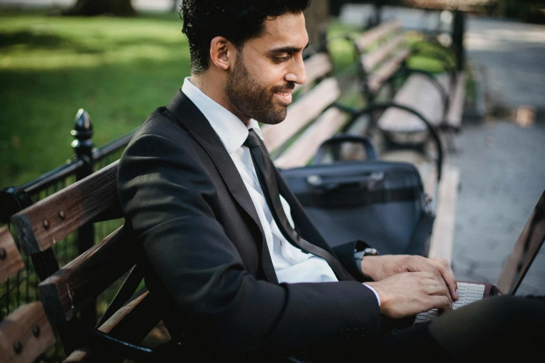 a man in a suit is sitting on a park bench using his laptop