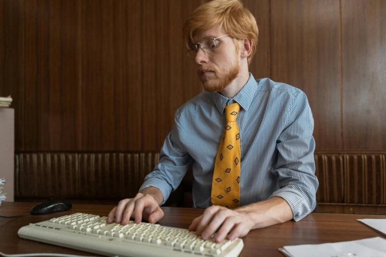 a man with glasses and a shirt and yellow tie sits at a keyboard in front of a monitor