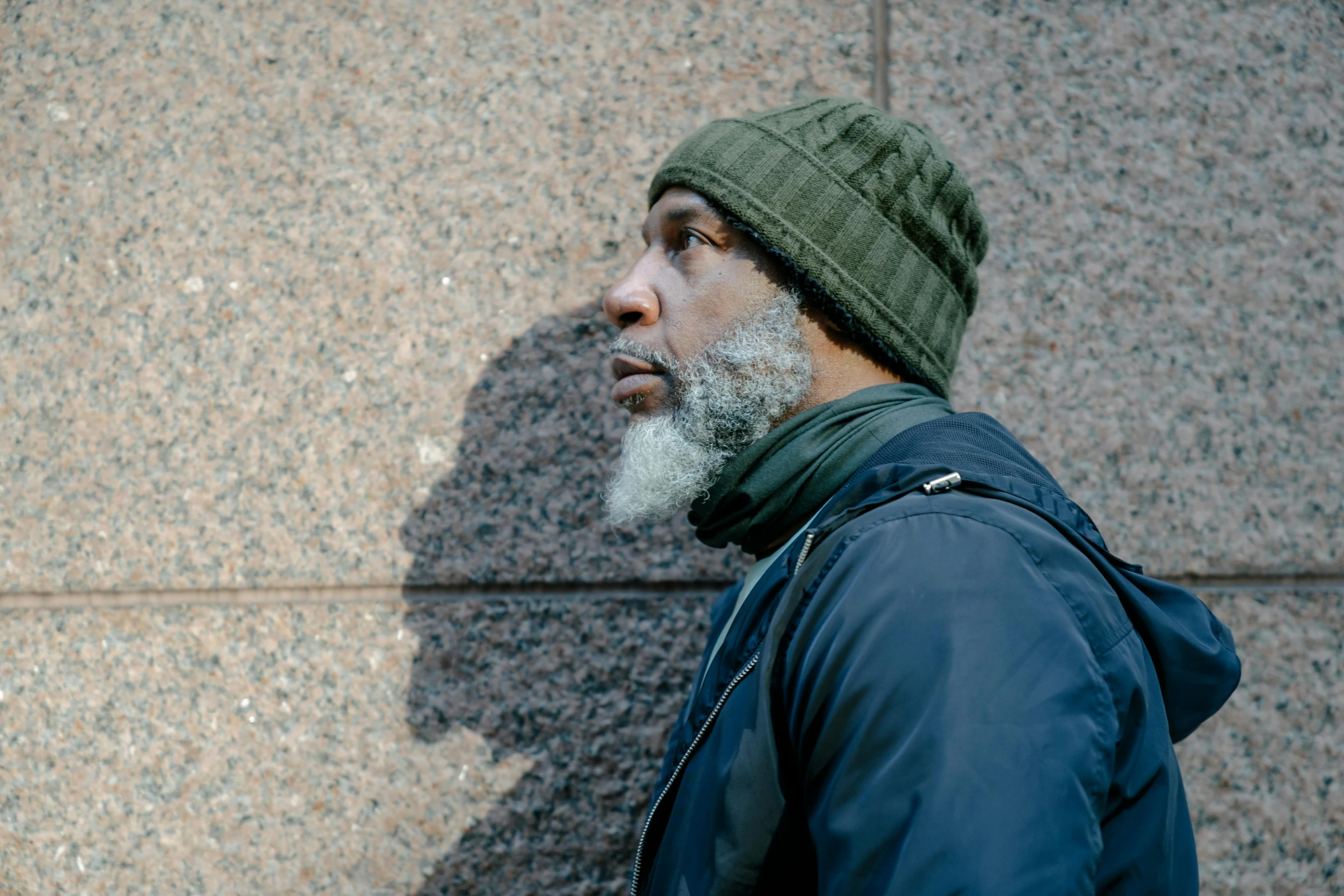 a bearded man wearing a cap and jacket standing in front of a wall