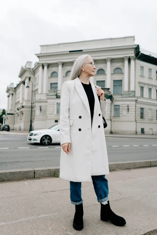 a woman with grey hair in a coat and booties stands on the street corner