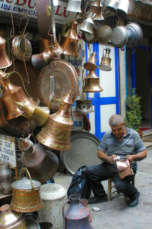 an old man sitting on a chair in front of various copper pots and pans