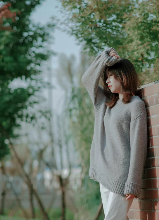 the young woman in white pants and sweater stands near a brick wall