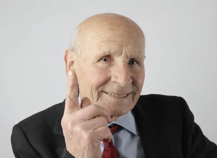 an elderly man in suit and tie holding up his finger to his face