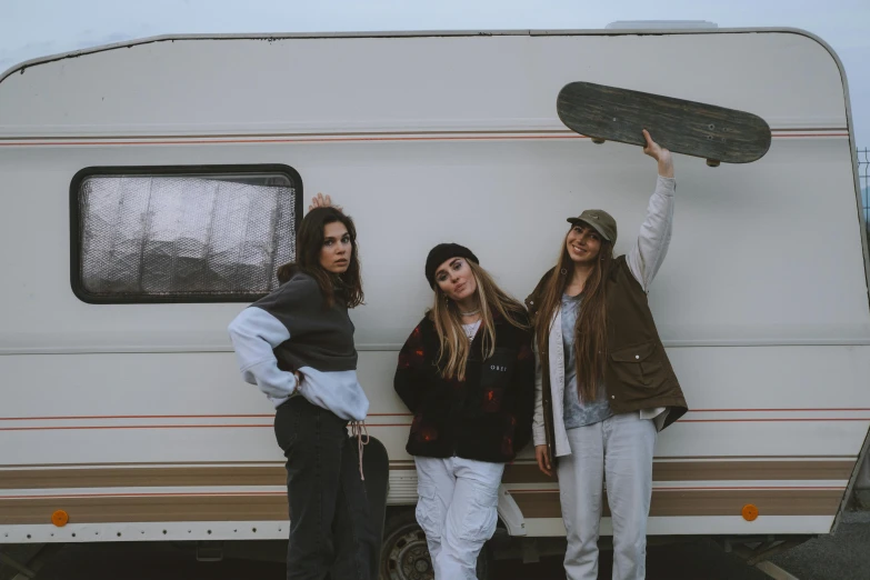 three girls standing next to an rv holding up a skateboard