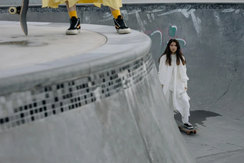 a  wearing sneakers stands at the edge of an indoor skate park with her skateboard as well