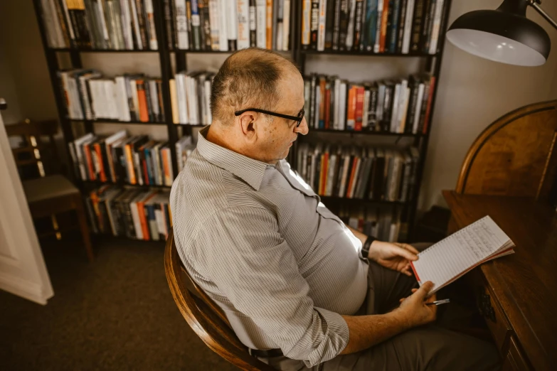a man in glasses sits at a desk while reading
