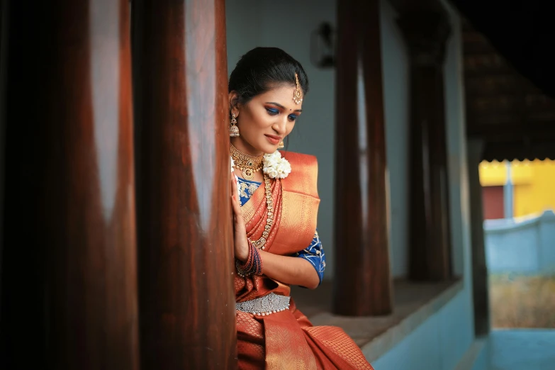 a woman wearing an orange sari, standing behind columns and holding a large vase