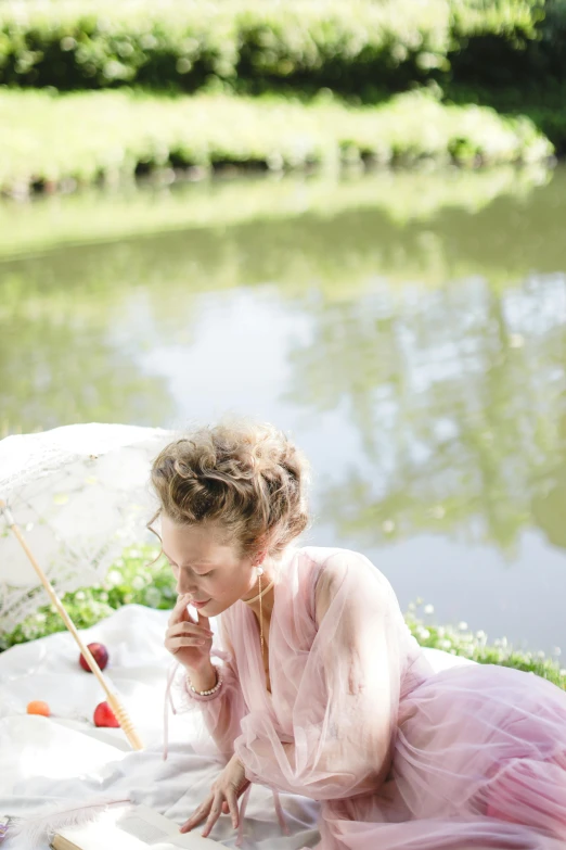 a girl wearing a pink dress and sitting on the ground near the water with an umbrella