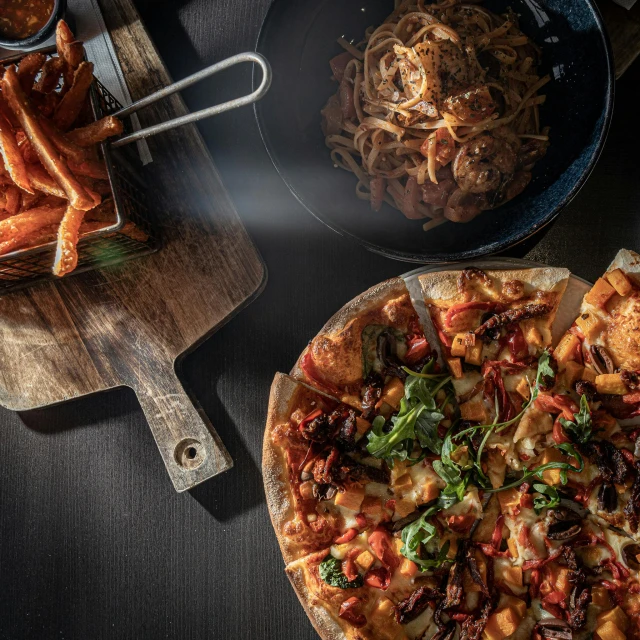 an uncooked pizza sits in front of a tray of pasta, with a pair of utensils on the table