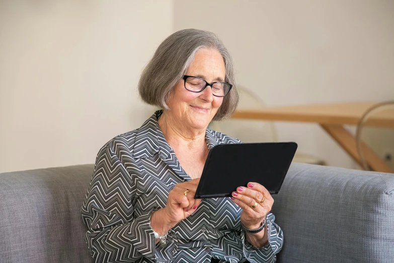 an old woman sitting on a gray couch, using her tablet