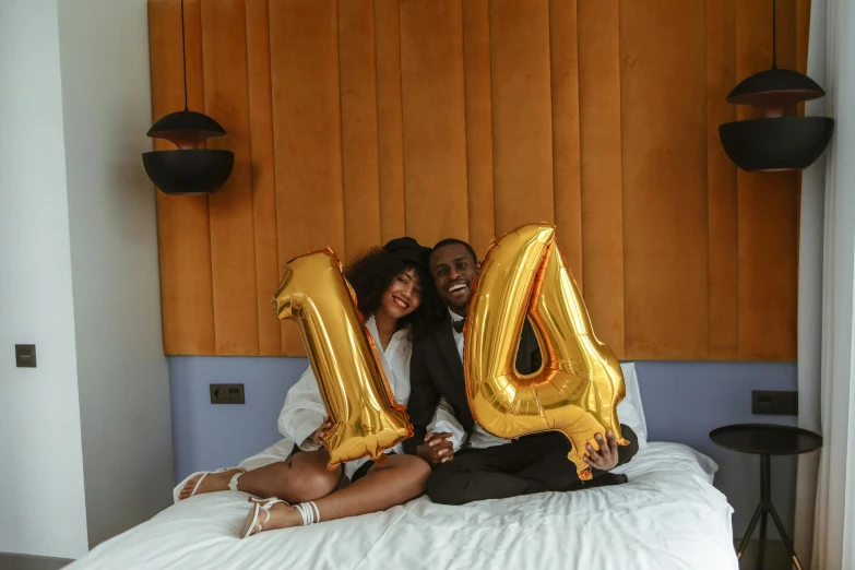 a couple sitting on top of a bed holding large golden balloons