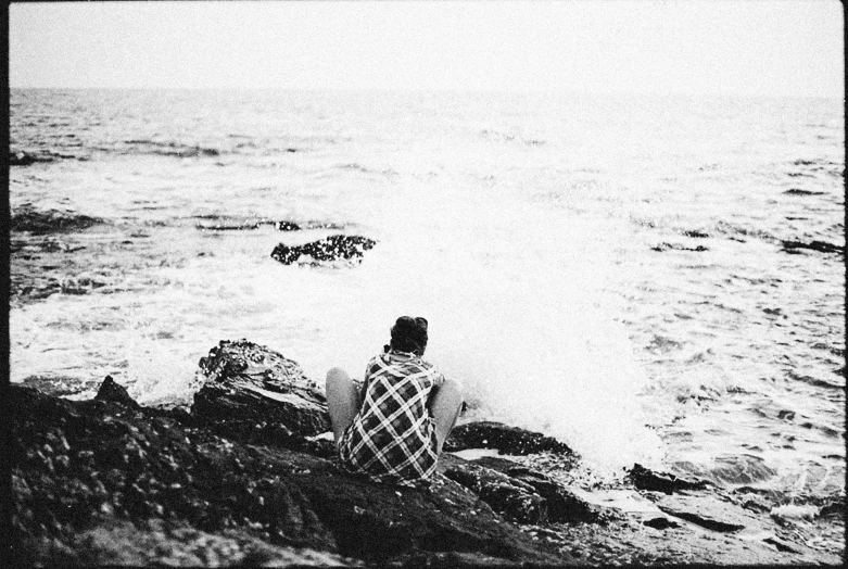 person sitting on rocky outcropping by ocean with surfboards