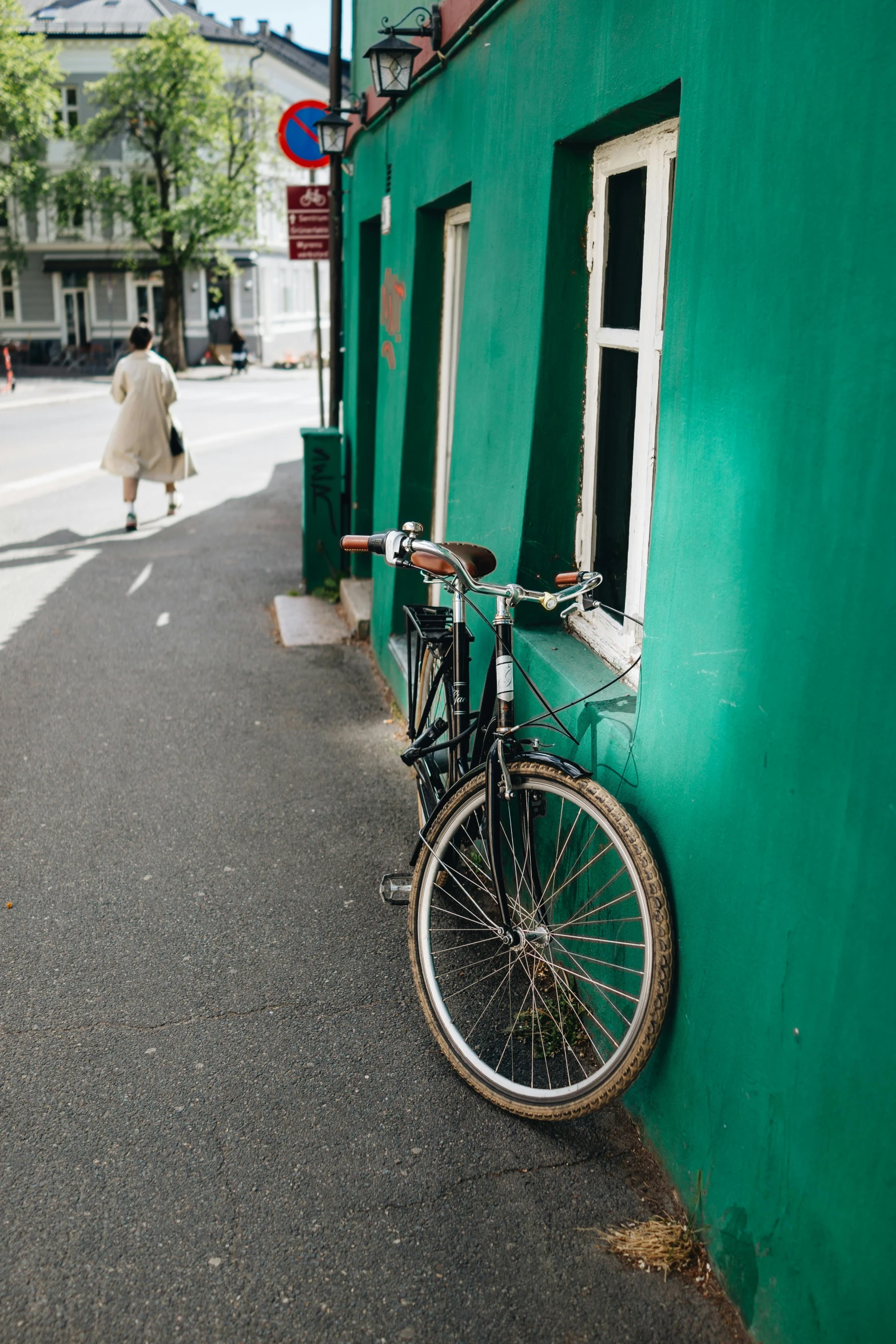 a black bicycle leaning against a green building with a person walking along the street