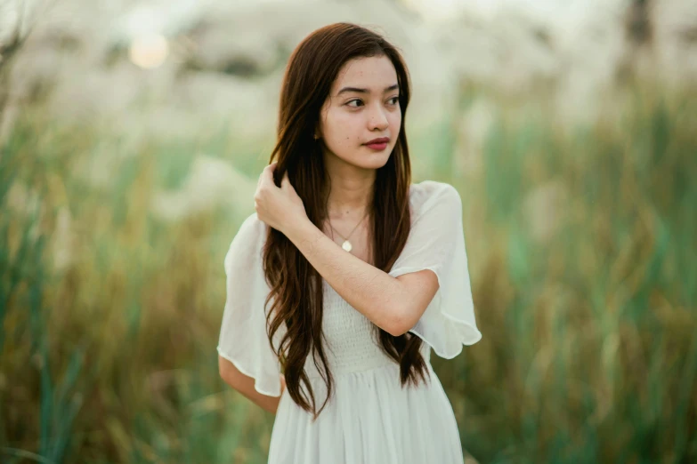  in white dress standing in grass with hand near her ear