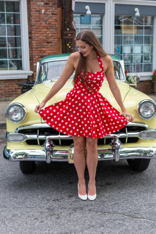 a woman in a polka dotted dress leans on the hood of an old yellow car