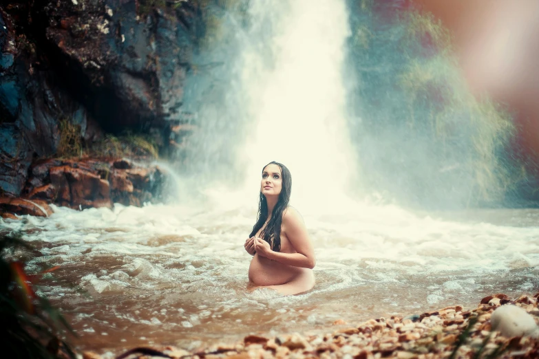 a woman with long hair wearing a wet suit sits in water near a waterfall