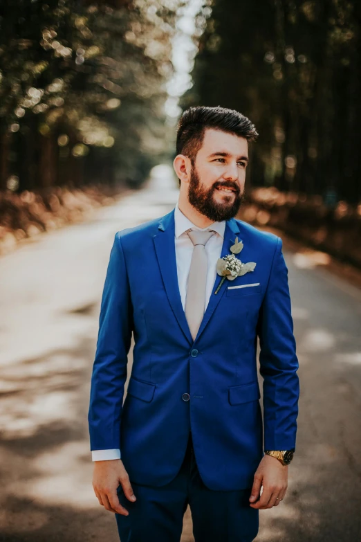 man with beard wearing a blue suit and tie stands in the middle of a tree lined road