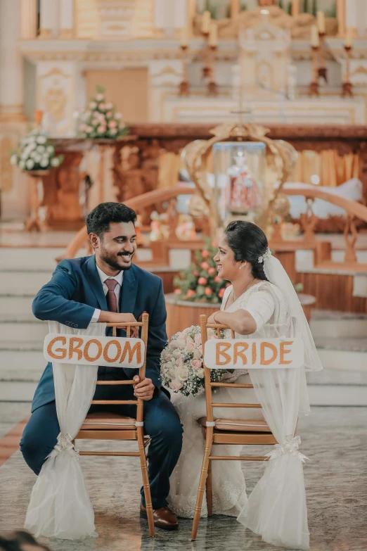 two brides in blue sitting on chairs holding up a sign that says groom and bride