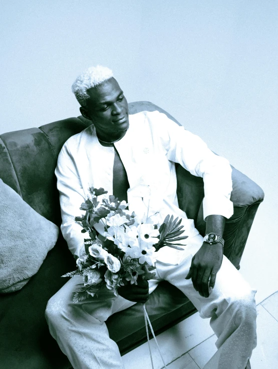 a black man with a white dress coat on sitting on a couch with flowers