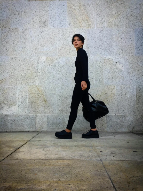 a woman walking down a sidewalk with a black bag on her shoulder