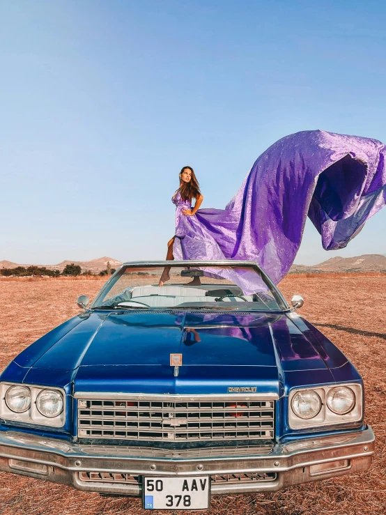 a woman stands on top of a blue convertible car in an arid area