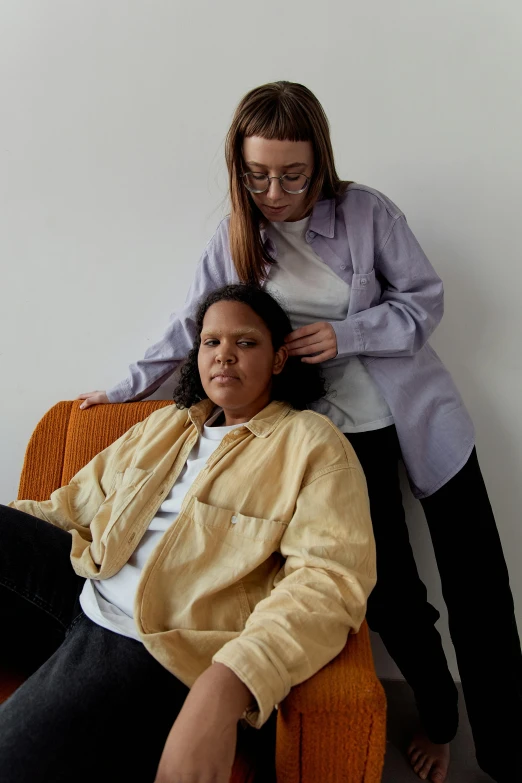 two women standing in front of the back of a chair with a person sitting next to them on a bed