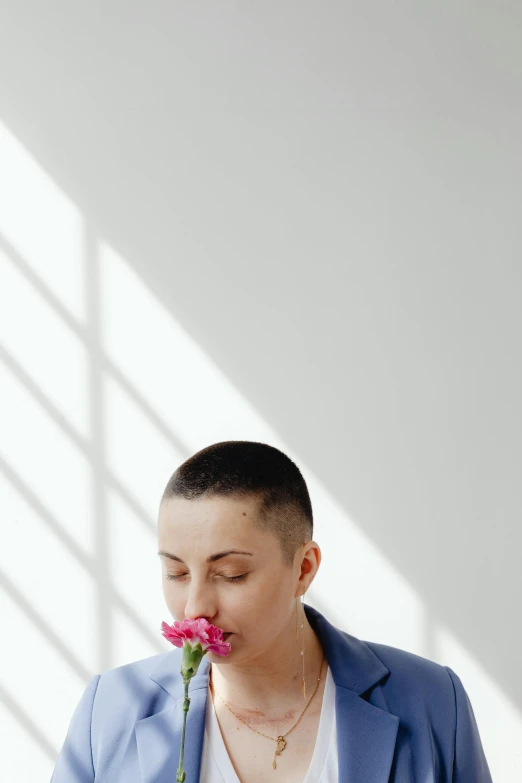 a woman with short hair, wearing blue jacket and a necklace holds a pink flower to her mouth and poses