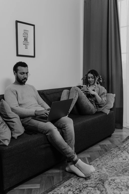 a man and woman sit on a couch using laptops