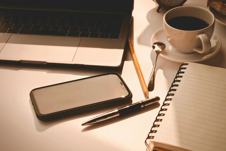 a laptop and pen, mug, and empty notepad on a white desk