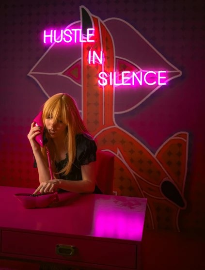 woman sitting on desk with phone in front of pink neon sign