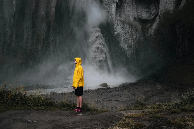 a person wearing yellow coat and black shorts stands in front of waterfall