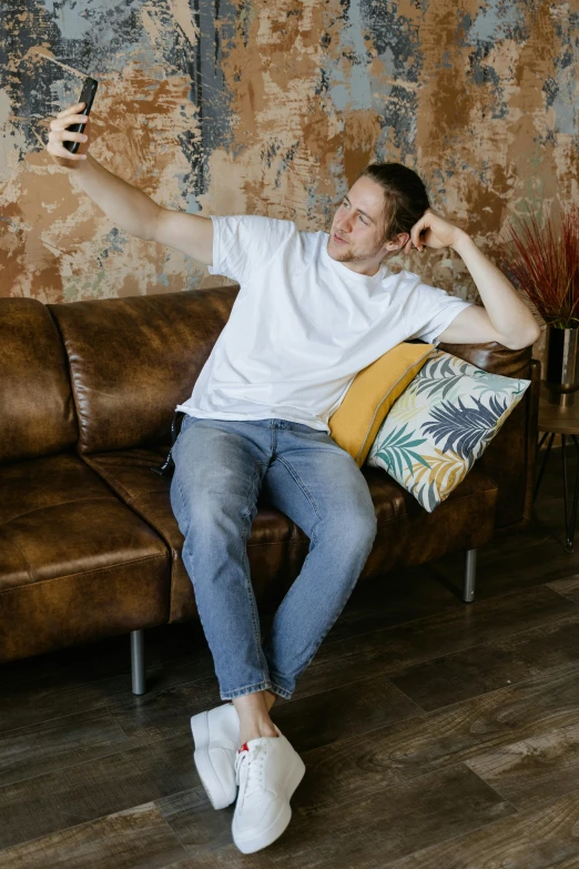 a man sitting on a brown couch with two pillows and his cellphone