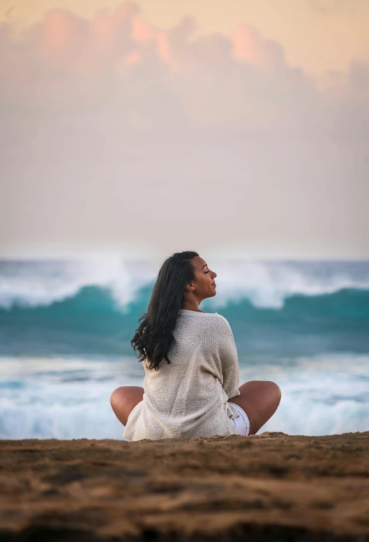 a woman in a sweater is sitting on the beach with her knees crossed