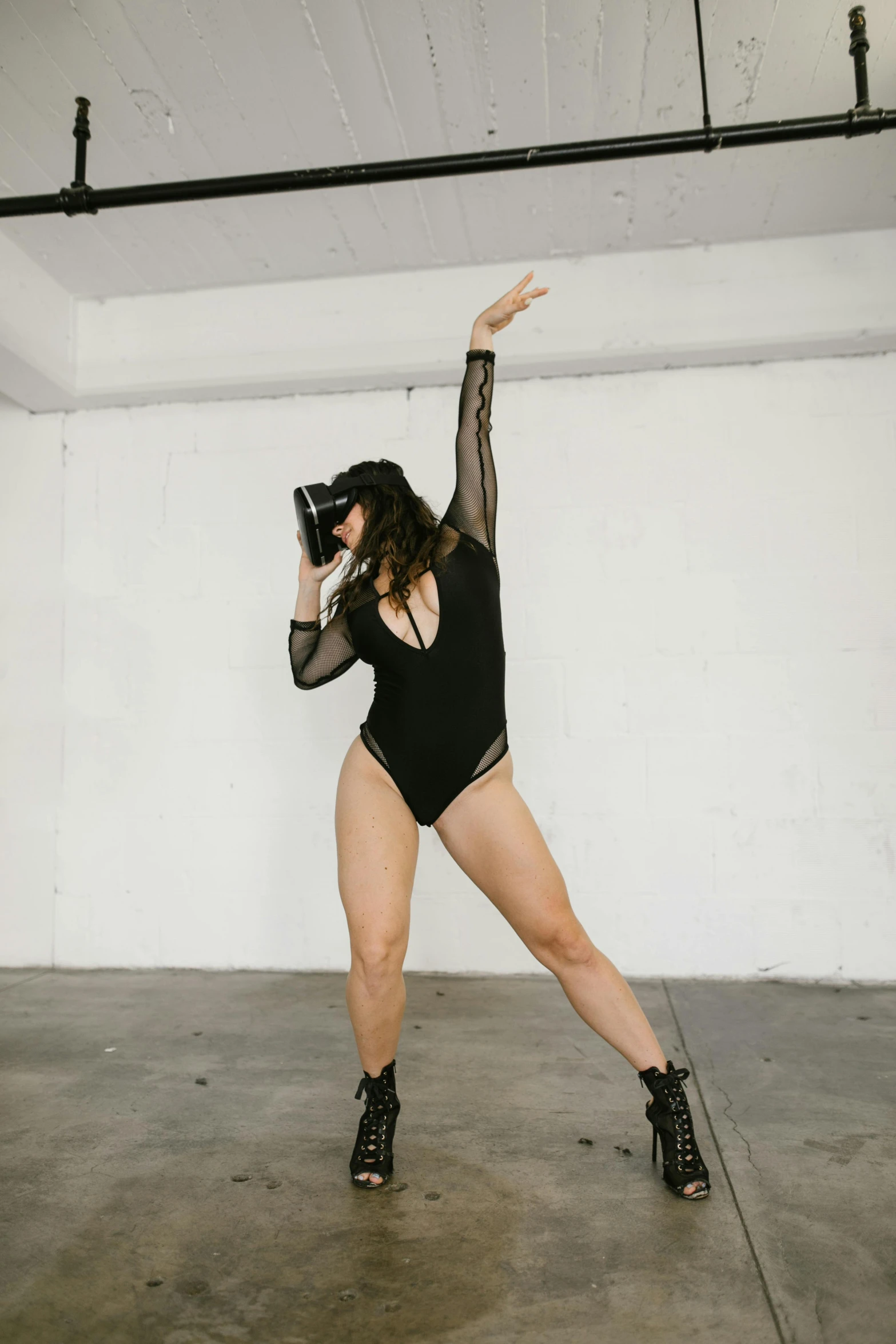 a woman wearing a one piece bodysuit and high heel sandals poses in the middle of a studio with her arms outstretched