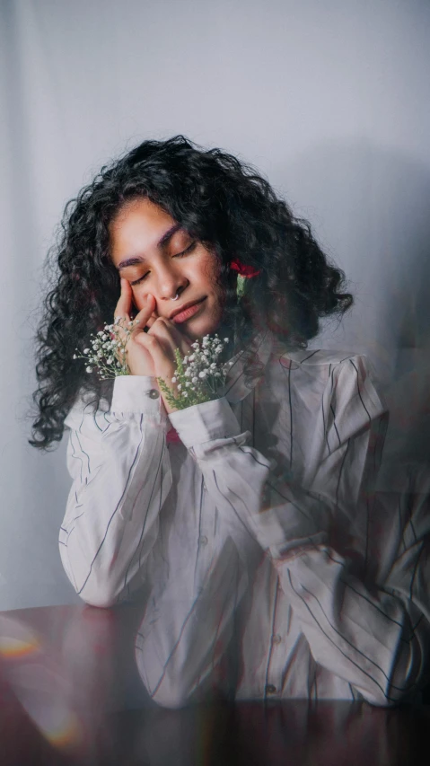 a young woman with curly hair wearing a white shirt holds her hand on her face